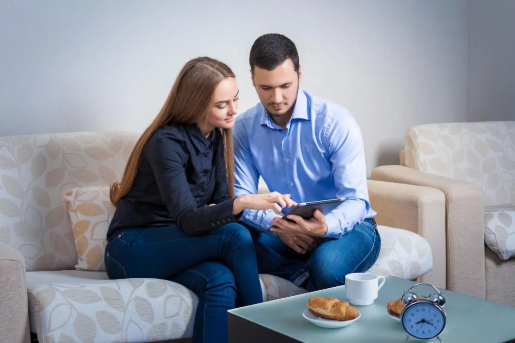 young-couple-laughing-sitting-sofa-sharing-information-displayed-electronic-tablet-with-each-other-drinking-coffee